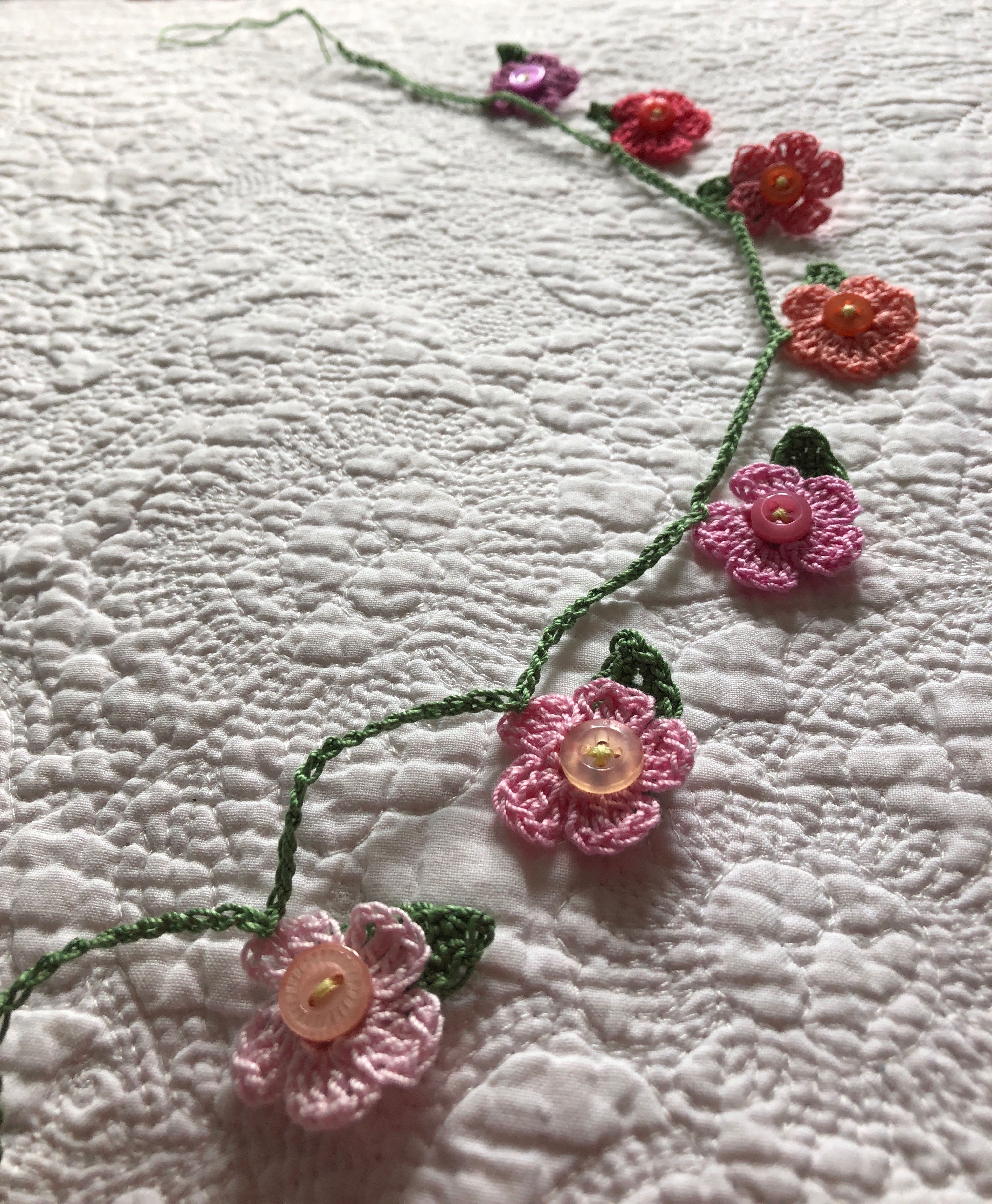 A handmade, crocheted garland of tiny flowers and leaves with a button detailing. The flowers are in a gradient of pastel shades from pale pink to purple.