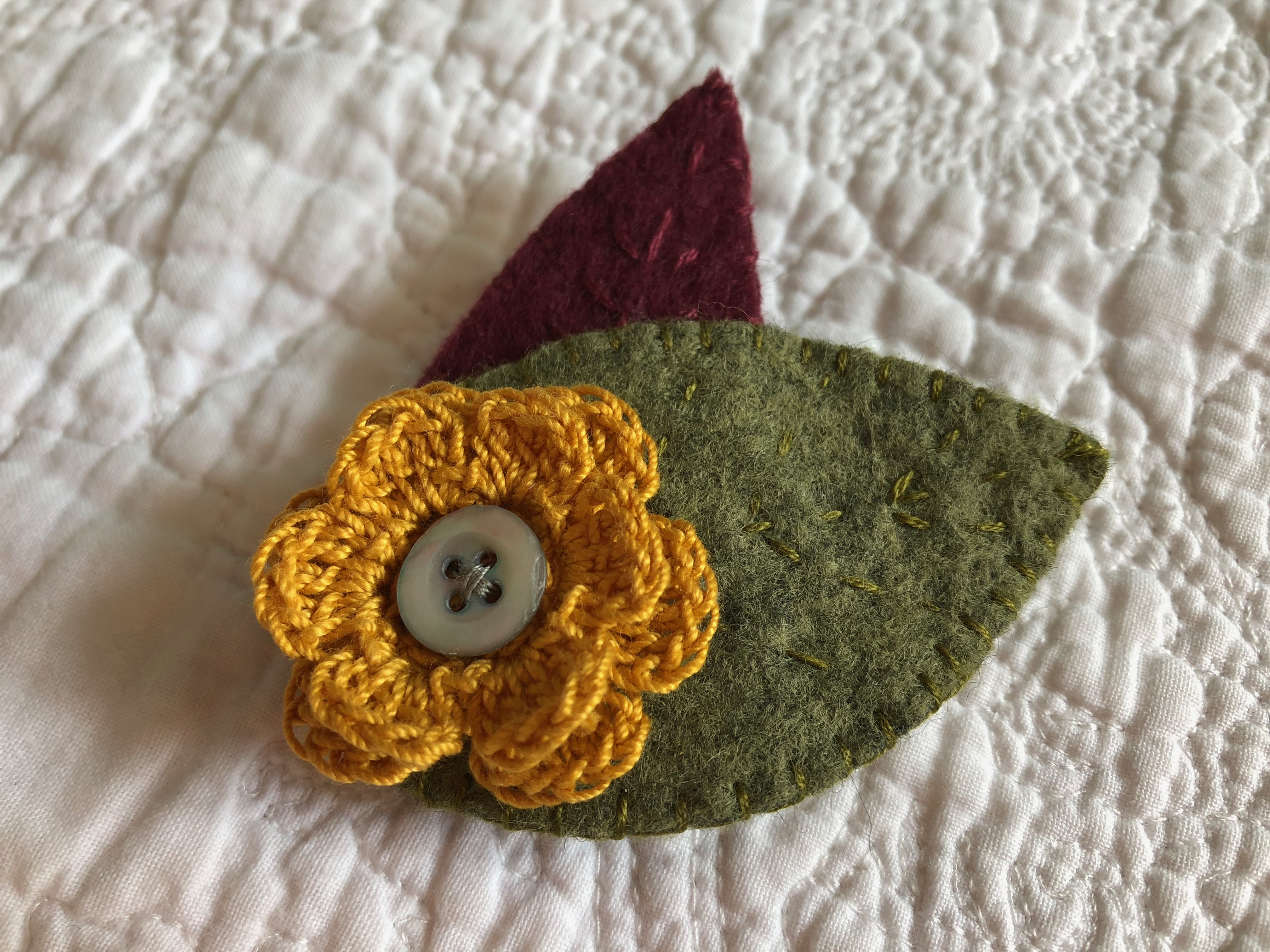 A handmade felt and crocheted brooch with hand stitched and button detailing. Mustard yellow flower with burgundy and green hand detailed felt leaves.