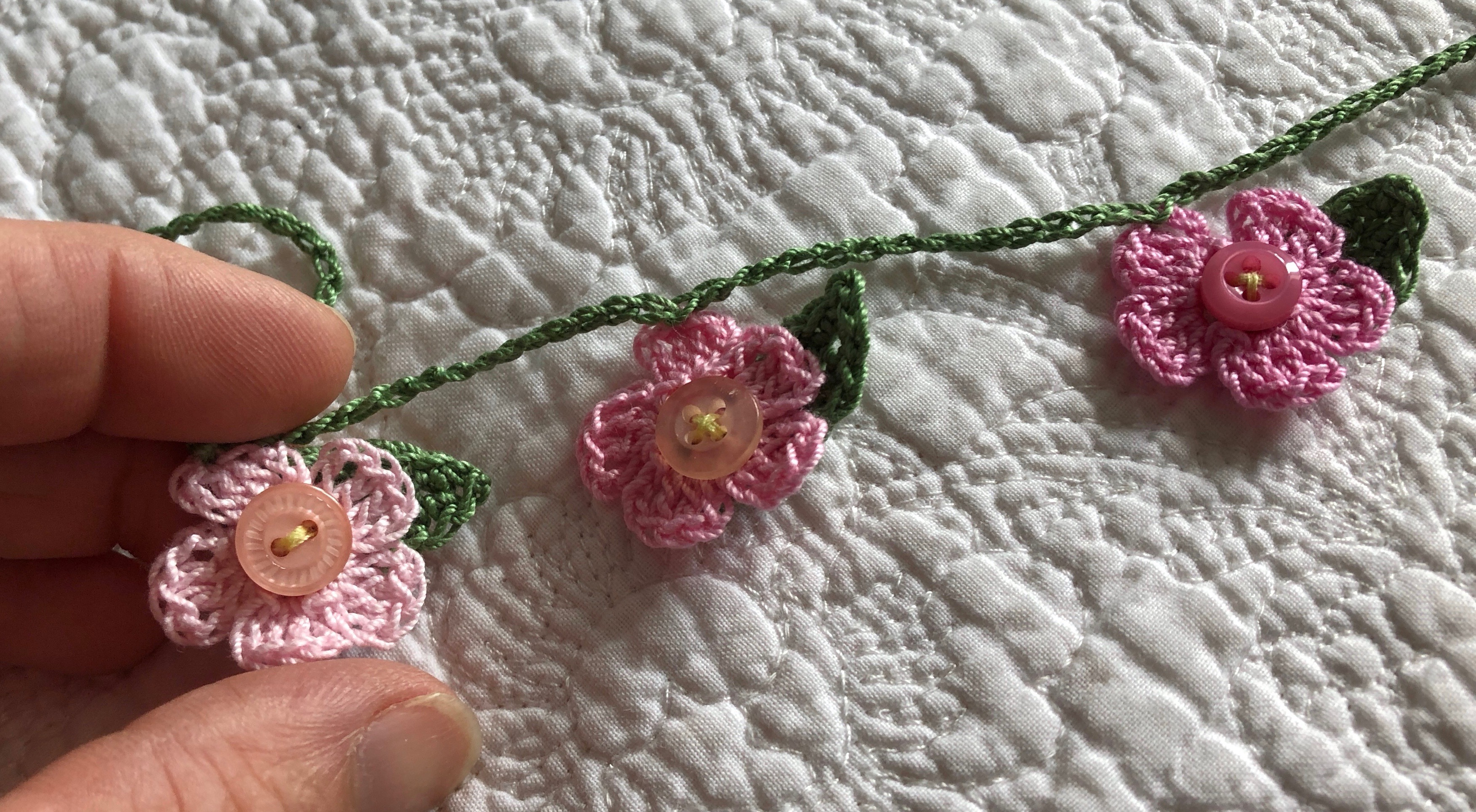 A handmade, crocheted garland of tiny flowers and leaves with a button detailing. The flowers are in a gradient of pastel shades from pale pink to purple.