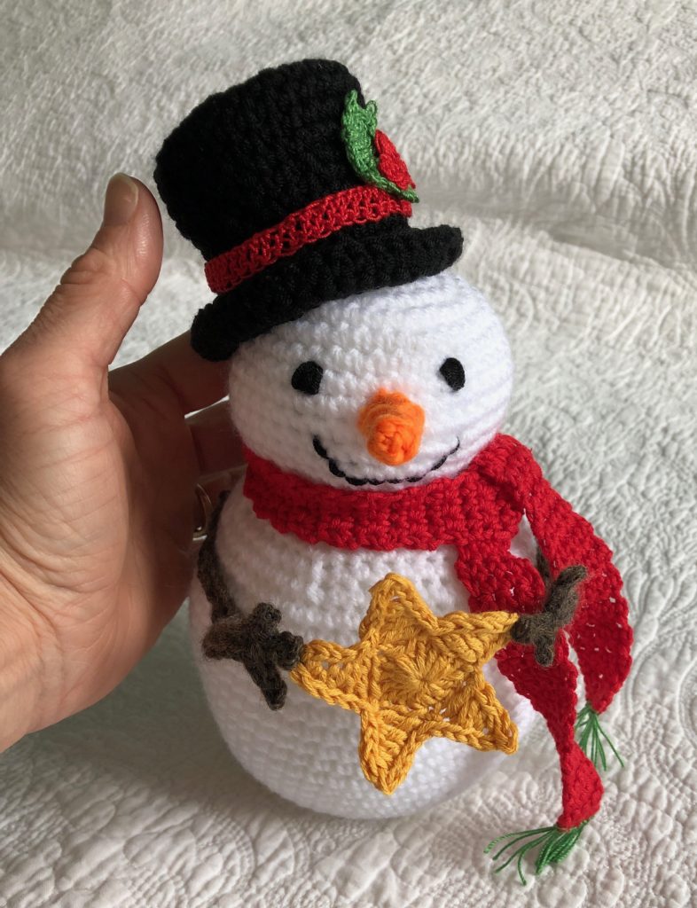 A crocheted snowman christmas tree topper wearing a black top hat with red band and green holly decoration, a red scarf and holding a yellow crocheted star.