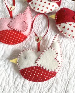 A hand cut and hand stitched, felt and fabric bird, with a red ribbon hanging loop and wooden heart button detail. Robins approximate size 8cm w x 5cm h. (excluding hanging ribbon.)