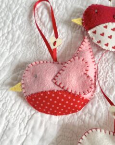 A hand cut and hand stitched, felt and fabric bird, with a red ribbon hanging loop and wooden heart button detail.