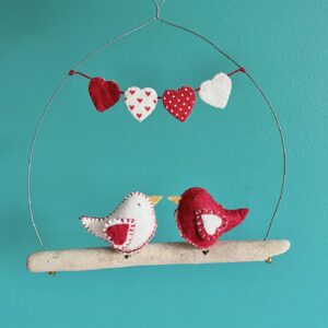 A pair of little love birds. Each one is handmade in felt with a red heart print, cotton fabric chest. These birds are sat on a natural driftwood perch with a wire hanger that is decorated with a garland of fabric and felt hearts. Approximate size 19.5cm width x 20cm height. This is a decoration only, and is NOT a toy. Keep out of the reach of babies and small children due to the small parts.