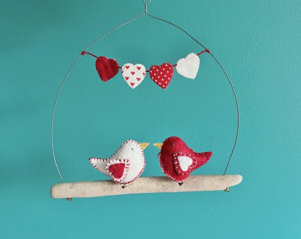 A pair of little love birds. Each one is handmade in felt with a red heart print, cotton fabric chest. These birds are sat on a natural driftwood perch with a wire hanger that is decorated with a garland of fabric and felt hearts. Approximate size 19.5cm width x 20cm height. This is a decoration only, and is NOT a toy. Keep out of the reach of babies and small children due to the small parts.