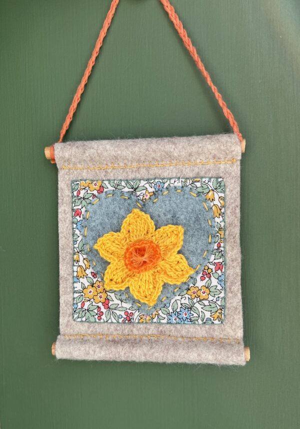 A small hand stitched decorative wall hanger, handmade using felt and 100% cotton fabric with a single crocheted Daffodil on a heart motif. The Daffodil and hanging loop are crocheted using 100% cotton. The piece is supported by 2 solid wooden dowels. The approximate measurements for this item are 9.5cm wide x 10.5cm Height x 0.8cm depth. (Not including the hanging cord.) This item is unique and handmade. No two pieces will be exactly the same. It has been designed and crafted as a hanging decoration and is not a toy. Keep away from babies and young children. Keep away from naked flames.