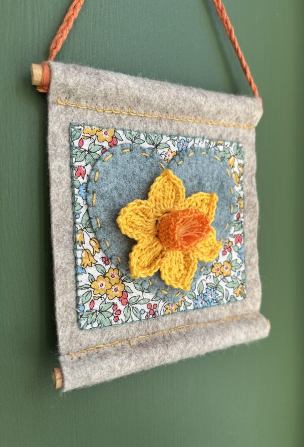 A small hand stitched decorative wall hanger, handmade using felt and 100% cotton fabric with a single crocheted Daffodil on a heart motif. The Daffodil and hanging loop are crocheted using 100% cotton. The piece is supported by 2 solid wooden dowels. The approximate measurements for this item are 9.5cm wide x 10.5cm Height x 0.8cm depth. (Not including the hanging cord.) This item is unique and handmade. No two pieces will be exactly the same. It has been designed and crafted as a hanging decoration and is not a toy. Keep away from babies and young children. Keep away from naked flames.