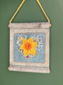 A small hand stitched decorative wall hanger, handmade using felt and 100% cotton fabric with a single crocheted Daffodil on a heart motif. The Daffodil and hanging loop are crocheted using 100% cotton. The piece is supported by 2 solid wooden dowels. The approximate measurements for this item are 10cm wide x 11.5cm Height x 0.8cm depth. (Not including the hanging cord.) This item is unique and handmade. No two pieces will be exactly the same. It has been designed and crafted as a hanging decoration and is not a toy. Keep away from babies and young children. Keep away from naked flames.