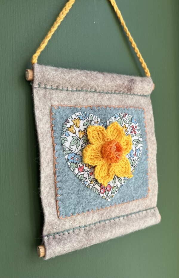 A small hand stitched decorative wall hanger, handmade using felt and 100% cotton fabric with a single crocheted Daffodil on a heart motif. The Daffodil and hanging loop are crocheted using 100% cotton. The piece is supported by 2 solid wooden dowels. The approximate measurements for this item are 10cm wide x 11.5cm Height x 0.8cm depth. (Not including the hanging cord.) This item is unique and handmade. No two pieces will be exactly the same. It has been designed and crafted as a hanging decoration and is not a toy. Keep away from babies and young children. Keep away from naked flames.