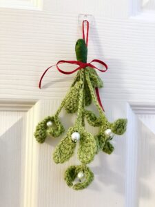 A handmade, crocheted, hanging mistletoe decoration. Made using green yarn and glass beads which are grouped and wired together and tied with a red ribbon to hang. Small size approximately 22cm high (not including ribbon hanging loop.) 5 springs of mistletoe are used in this arrangement.