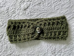 A hand crocheted, textured, ear warmer, in a warm green colour, with button detail. Made using a super soft, warm and chunky Acrylic 75% / Wool 25% mix yarn.