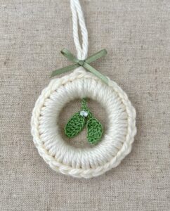 A crocheted and mini mistletoe embellished Christmas wreath, hanging decoration. Handmade using a wooden ring with cream coloured yarn, 100% cotton in green and opal effect glass beaded embellishments. Approximate size of wreath 8.5cm height x 7cm width. Not including hanging loop.