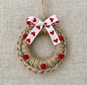 A crocheted and mini embellished Christmas wreath, hanging decoration. Handmade using a wooden ring with 100% natural coloured Jute and red glass beaded embellishments, with a heart detail bow. Approximate size of wreath 8.5cm height x 7cm width. Not including hanging loop.