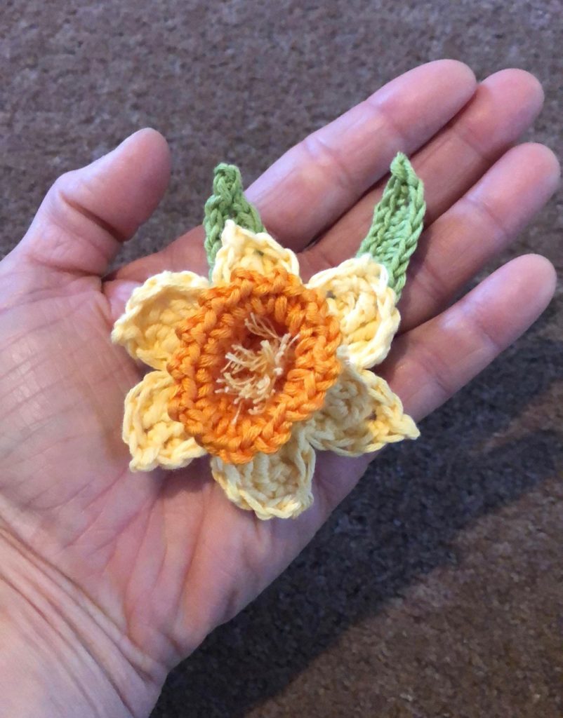 Crocheted yellow daffodil brooch with green leaves.