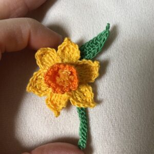 A crocheted, yellow daffodil flower with orange centre, green stem and leaf detail brooch. this has a metal locking pin fixing on the back. Made using 100% cotton.