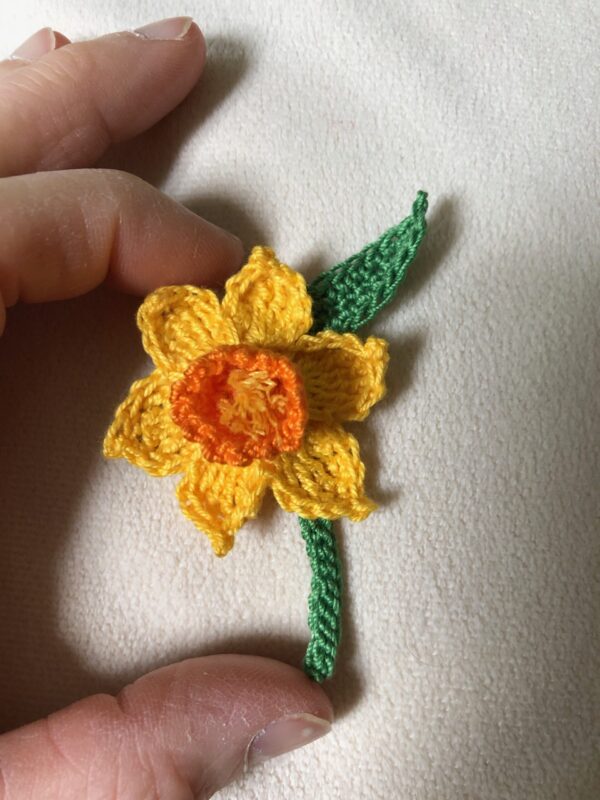 A crocheted, yellow daffodil flower with orange centre, green stem and leaf detail brooch. this has a metal locking pin fixing on the back. Made using 100% cotton.