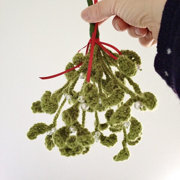 A handmade, crocheted, hanging mistletoe decoration. Made using green yarn and glass beads which are grouped and wired together and tied with a red ribbon to hang. Large size approximately 25cm high (not including ribbon hanging loop.) 12 springs of mistletoe are used in this arrangement.