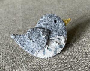 This little birdie brooch is completely hand cut, stitched, embroidered and embellished. It is made using a wool mix felt in a pale grey colour with a mistletoe patterned fabric chest. It is hand embellished using glass beads and has a metal locking fixing on the back.