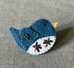 This little birdie brooch is completely hand cut, stitched, embroidered and embellished. It is made using a wool mix felt in a teal colour with a holly patterned fabric chest. It is hand embellished using glass beads and has a metal locking fixing on the back.