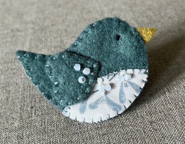 This little birdie brooch is completely hand cut, stitched, embroidered and embellished. It is made using a wool mix felt in a dark green colour with a mistletoe patterned fabric chest. It is hand embellished using glass beads and has a metal locking fixing on the back.