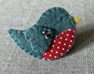 This little birdie brooch is completely hand cut, stitched, embroidered and embellished. It is made using a wool mix felt in a dark green colour with a spotty patterned fabric chest. It is hand embellished using glass beads and has a metal locking fixing on the back.