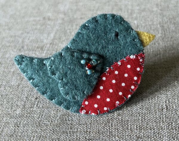 This little birdie brooch is completely hand cut, stitched, embroidered and embellished. It is made using a wool mix felt in a dark green colour with a spotty patterned fabric chest. It is hand embellished using glass beads and has a metal locking fixing on the back.