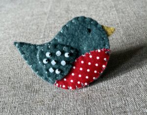 This little birdie brooch is completely hand cut, stitched, embroidered and embellished. It is made using a wool mix felt in a dark green colour with a spotty patterned fabric chest. It is hand embellished using glass beads and has a metal locking fixing on the back.