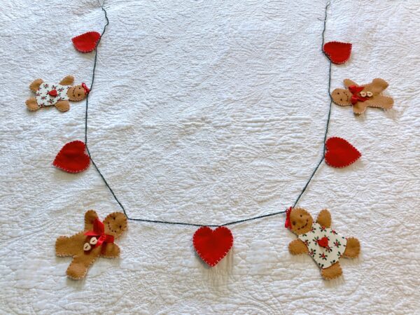 A garland of 4 gingerbread people and 5 red hearts. Hand-cut and handmade little gingerbread people and hearts, all made in felt, with fabric, glass beads, buttons and ribbon details, which are crocheted on to a cotton string. Approximate total length 125cm.