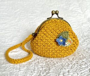 A small handmade and crocheted handheld purse. 
Made using 100% cotton yarn and embellished with a cotton and felt flower and leaf detail. 
Fitted with a solid, bronze coloured metal clasp closure, and a removable, crocheted wrist strap with brass coloured beads and clip.
