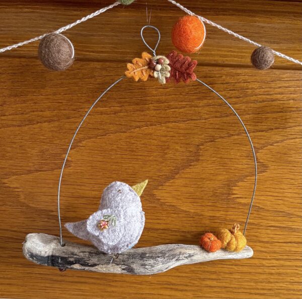 A single, small sized bird, handmade using a light mushroom coloured felt with a multi coloured floral print, cotton fabric chest. The bird is sat on a natural driftwood perch with a collection of little felt pumpkins. It's suspended on a wire hanger that is decorated with little felt autumn leaves and wooden beads. Approximate size 16cm width x 15.5cm height.