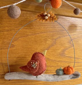 A single, small sized bird, handmade using a rust brown coloured felt with a multi coloured floral print, cotton fabric chest. The bird is sat on a natural driftwood perch with a collection of little felt pumpkins. It's suspended on a wire hanger that is decorated with little felt autumn leaves and wooden beads. Approximate size 15cm width x 15cm height.