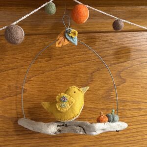 A single, small sized bird, handmade using a yellow coloured felt with a multi coloured floral print, cotton fabric chest. The bird is sat on a natural driftwood perch with a collection of little felt pumpkins. It's suspended on a wire hanger that is decorated with little felt autumn leaves and glass beads. Approximate size 14cm width x 14cm height.