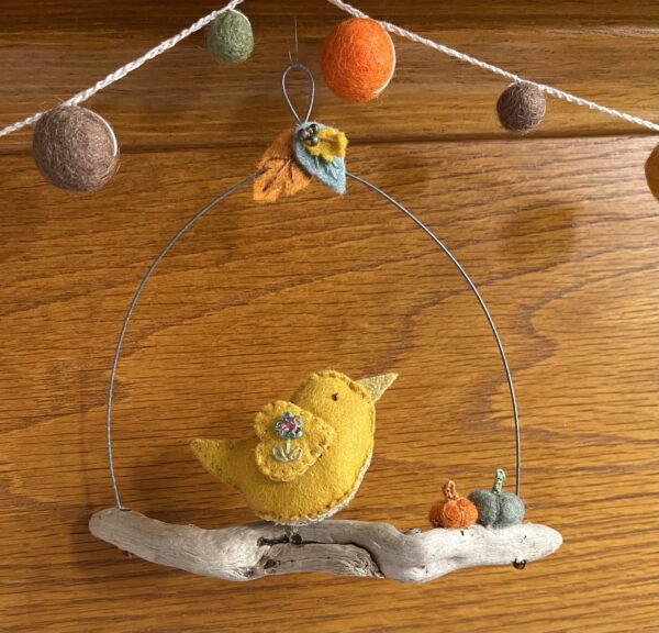 A single, small sized bird, handmade using a yellow coloured felt with a multi coloured floral print, cotton fabric chest. The bird is sat on a natural driftwood perch with a collection of little felt pumpkins. It's suspended on a wire hanger that is decorated with little felt autumn leaves and glass beads. Approximate size 14cm width x 14cm height.