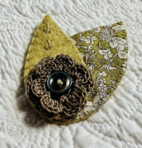 A single, double layered, crocheted flower in a coffee colour, with vintage button detail and two leaf brooch. Flower made using 100% Cotton with hand stitched felt and patterned fabric leaves. A locking metal brooch fastening on the back. Approximate size 7.5cm width x 6.5cm height.