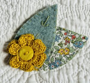 A single, double layered, crocheted flower in a mustard colour, with vintage button detail and two leaf brooch. Flower made using 100% Cotton with hand stitched felt and patterned fabric leaves. A locking metal brooch fastening on the back. Approximate size 7.5cm width x 6.5cm height.