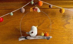 A single, small sized bird, handmade using a warm cream coloured felt with a multicoloured floral print, cotton fabric chest. The bird is sat on a natural driftwood perch with a collection of little felt pumpkins. It's suspended on a wire hanger that is decorated with little felt autumn leaves and wooden beads. Approximate size 15cm width x 16cm height.