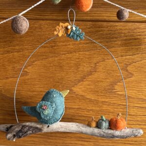 A single, small sized bird, handmade using a subtle green coloured felt with a multicoloured floral print, cotton fabric chest. The bird is sat on a natural driftwood perch with a collection of little felt pumpkins. It's suspended on a wire hanger that is decorated with little felt autumn leaves and wooden beads. Approximate size 19cm width x 16cm height.