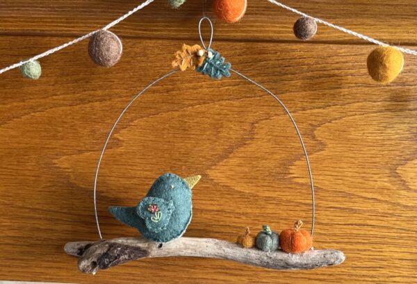 A single, small sized bird, handmade using a subtle green coloured felt with a multicoloured floral print, cotton fabric chest. The bird is sat on a natural driftwood perch with a collection of little felt pumpkins. It's suspended on a wire hanger that is decorated with little felt autumn leaves and wooden beads. Approximate size 19cm width x 16cm height.