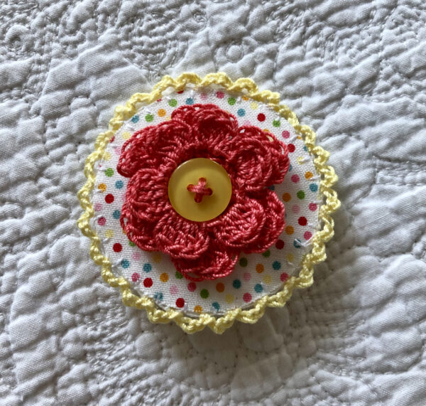 A round brooch made using a patterned fabric background with a crocheted flower in the centre and crocheted edging. Handmade using 100% cotton fabric and yarn. With button detail and a locking brooch fastening on a felt backing.